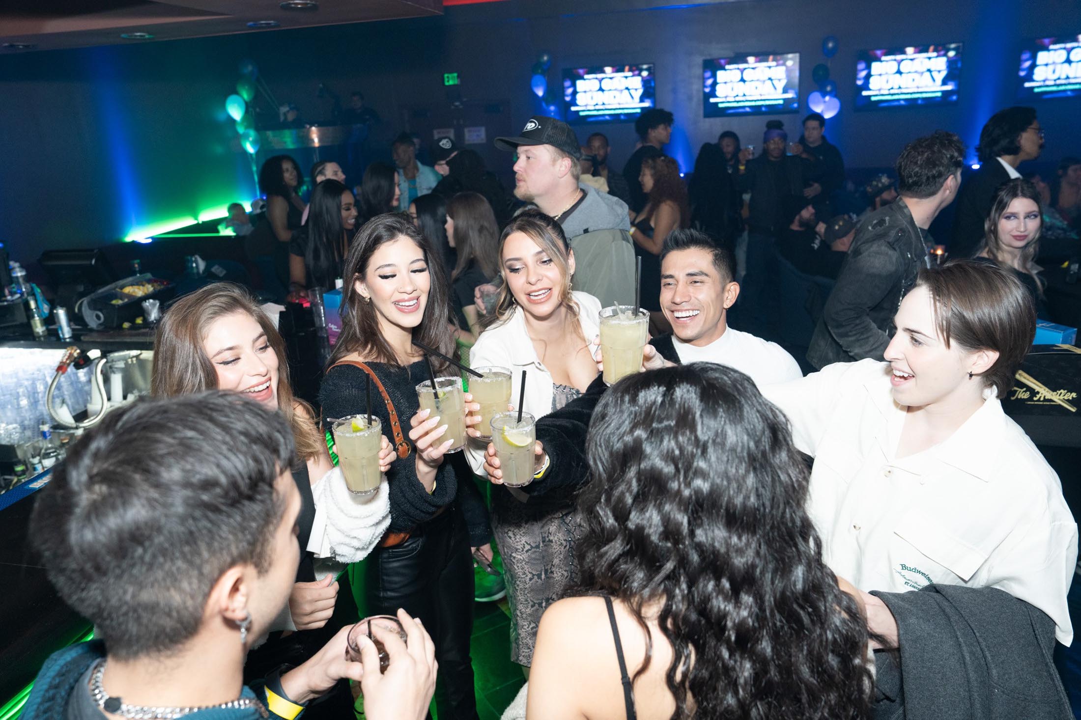 A group of people holding drinks and socializing in a bar with screens on the wall.